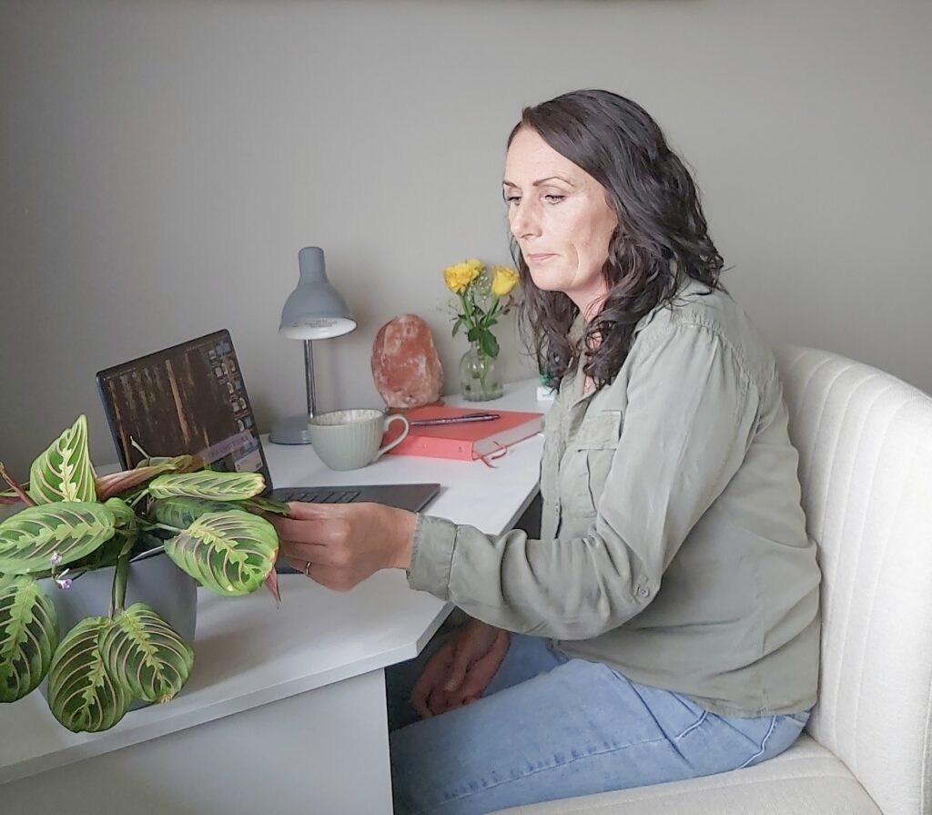 Client seated at a desk, gently touching a houseplant, while working on a laptop in natural light