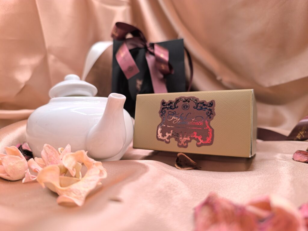 Luxury biscuit gift box beside a white teapot styled on soft pink fabric