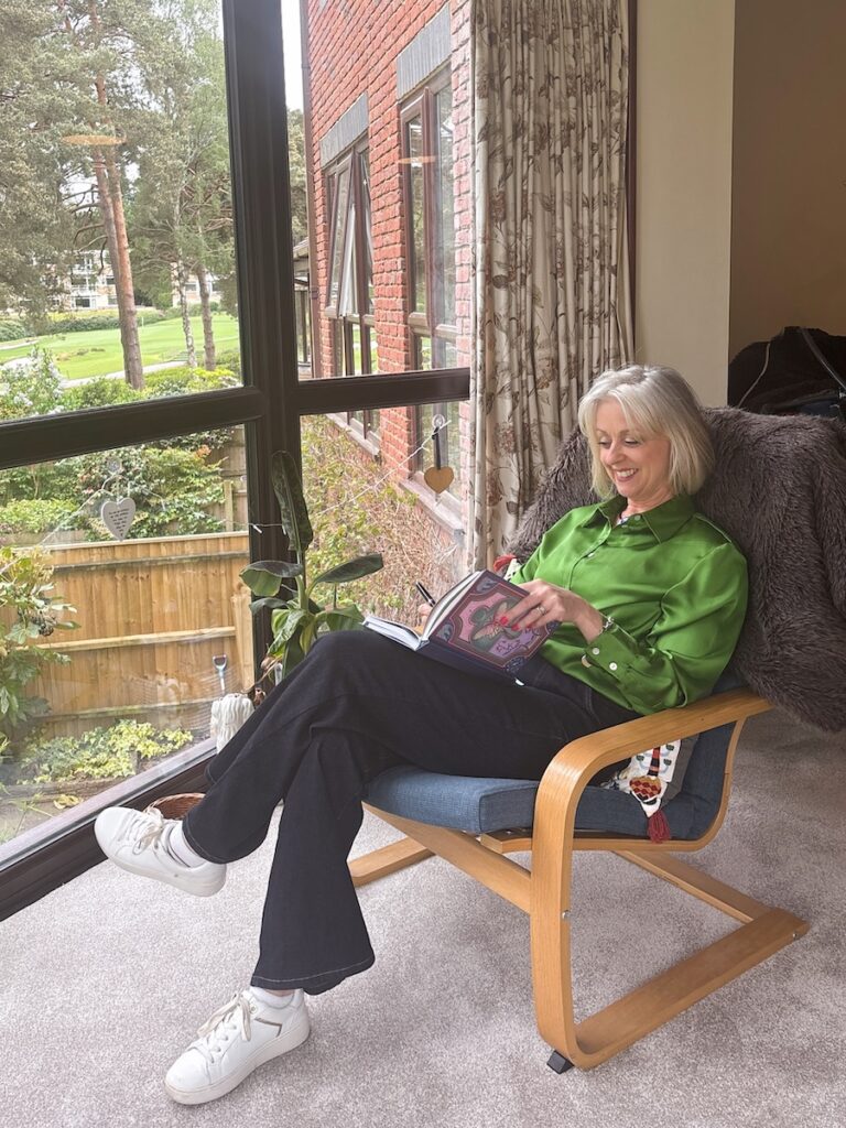 Jacqui sitting in a wooden armchair by a large window, smiling while planning an event in her notebook.
