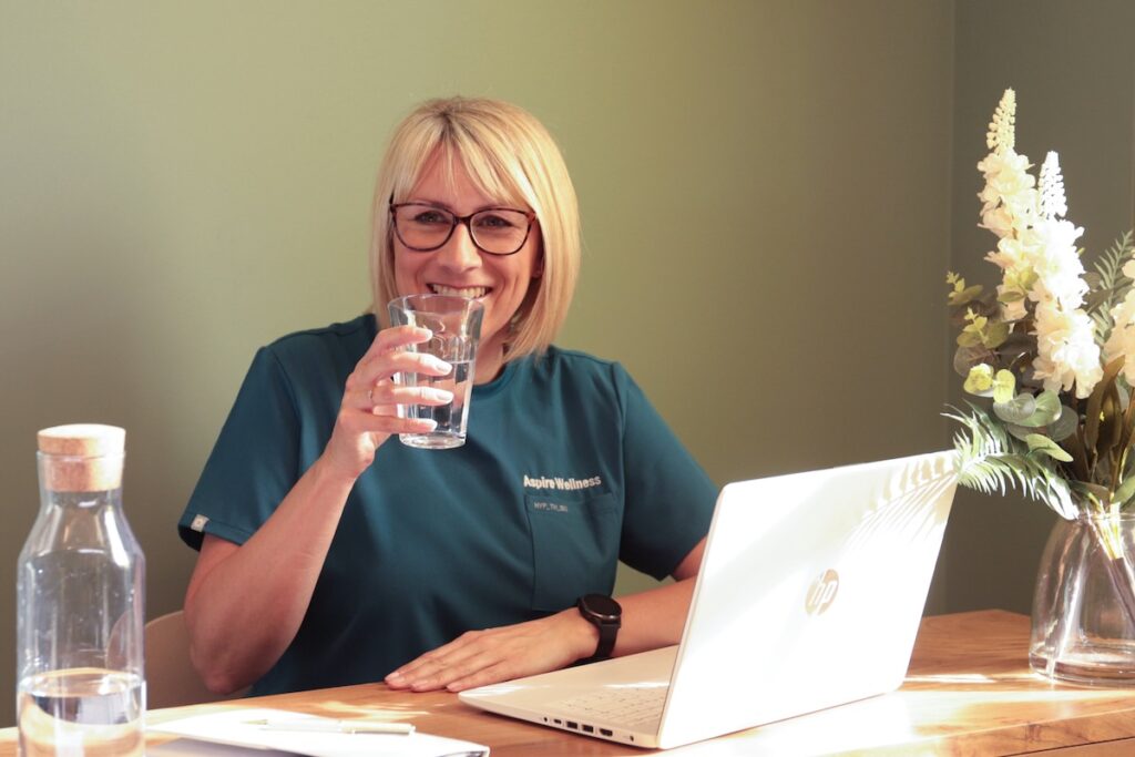 Kirsty smiling while holding a glass of water at a table with a laptop in a co-working space, wearing an Aspire Wellness uniform.