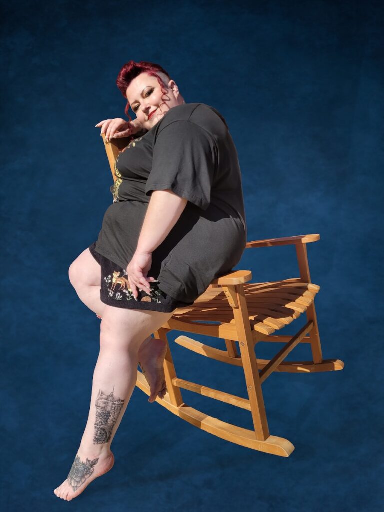 Lisa sitting sideways in a wooden rocking chair against a dark blue backdrop, smiling over her shoulder with her tattooed leg extended.