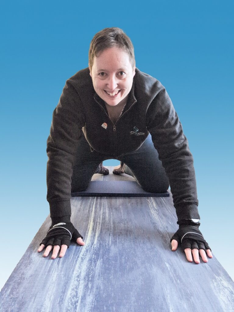 Lou smiling while holding a high plank position on an exercise mat, wearing black sportswear and fingerless gloves.