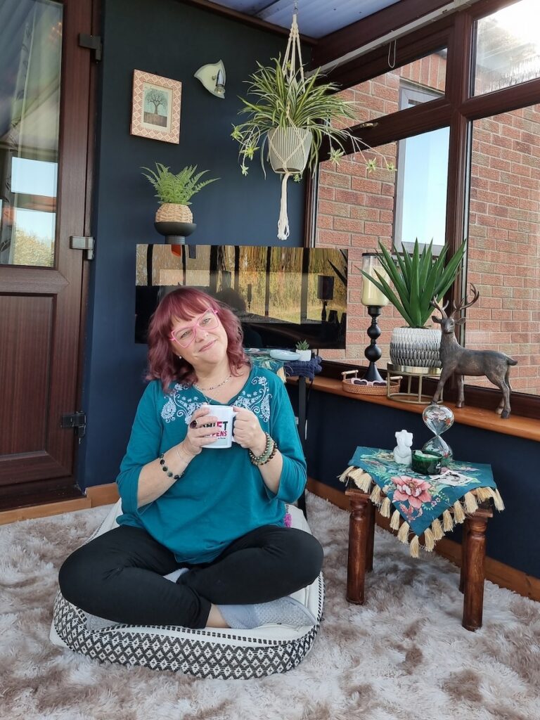 Nicky sitting cross-legged on a floor cushion holding a mug in her conservatory, with personal items displayed on a table beside her.