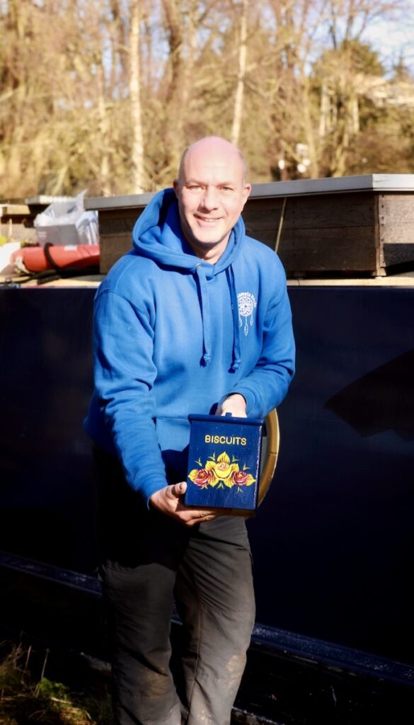 Paul smiling outdoors while holding a blue biscuit tin decorated with traditional canal roses and castles.