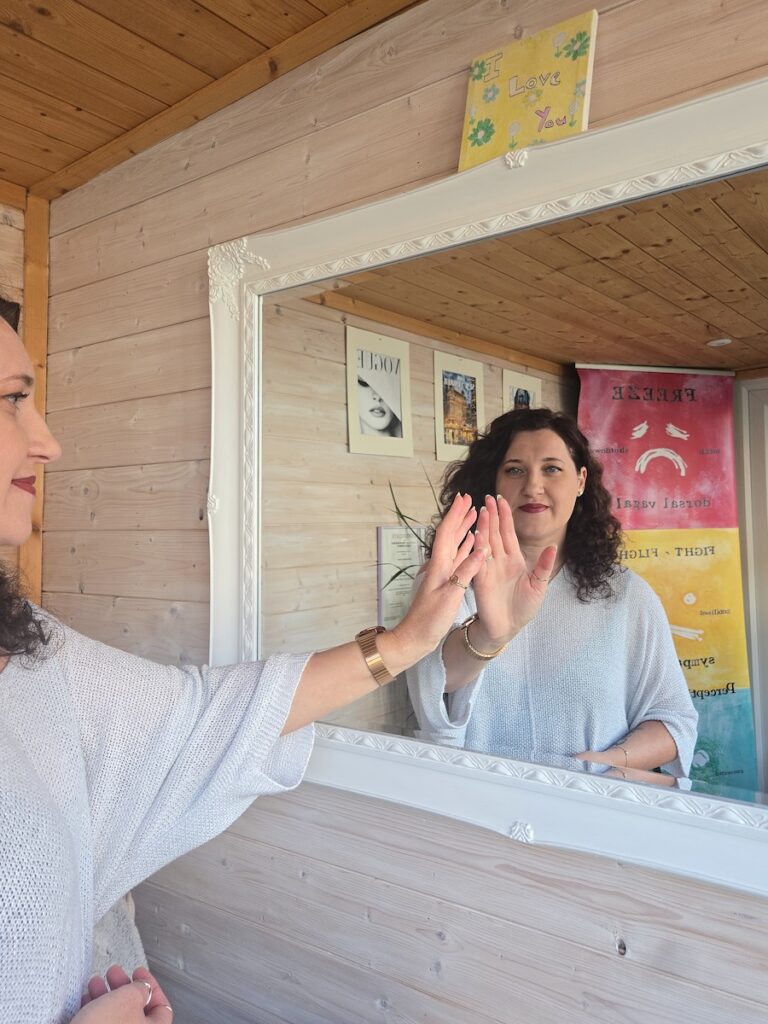 Ralu looking at her reflection in an ornate mirror while raising one hand to touch the glass in her wooden garden office.