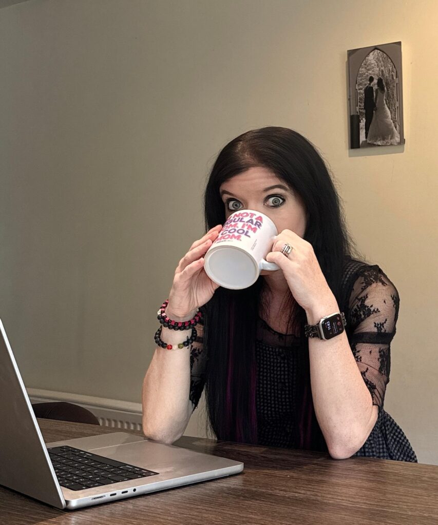 Sam smiling over a mug reading “Not a Regular Mum, a Cool Mum” while sitting beside a laptop during a virtual brand photo shoot.
