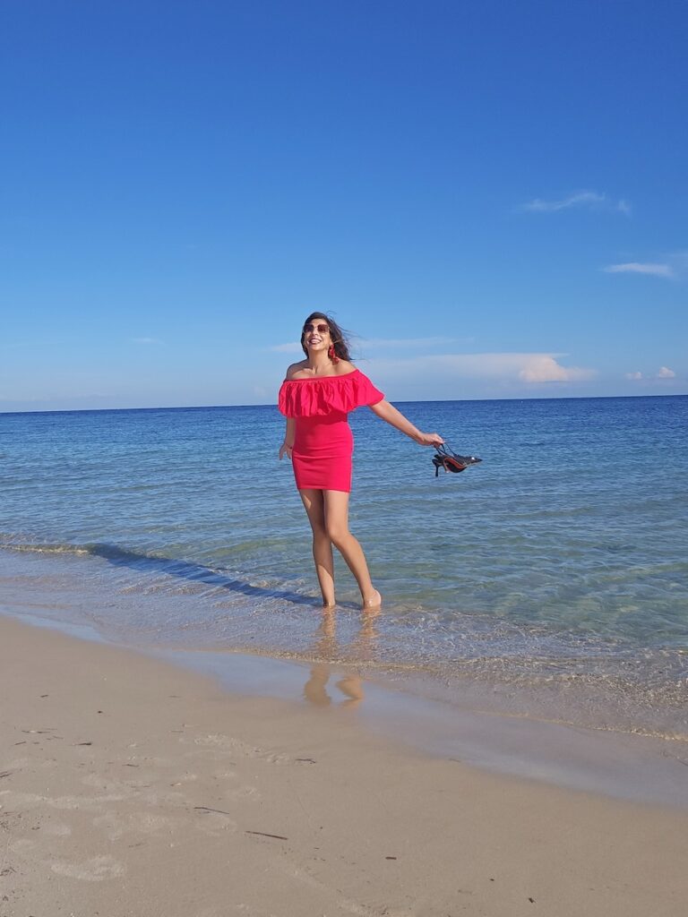 Sharan standing barefoot at the water’s edge on a beach in Spain, smiling towards the sky while holding her sandals and wearing a red off-the-shoulder dress.
